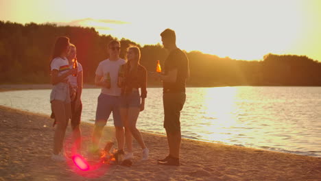 The-five-best-friends-spend-time-at-sunset-on-the-sand-beach-in-shorts-and-t-shirts-around-bonfire-with-beer.-They-are-talking-to-each-other-and-enjoying-the-summer-evening-in-nature.